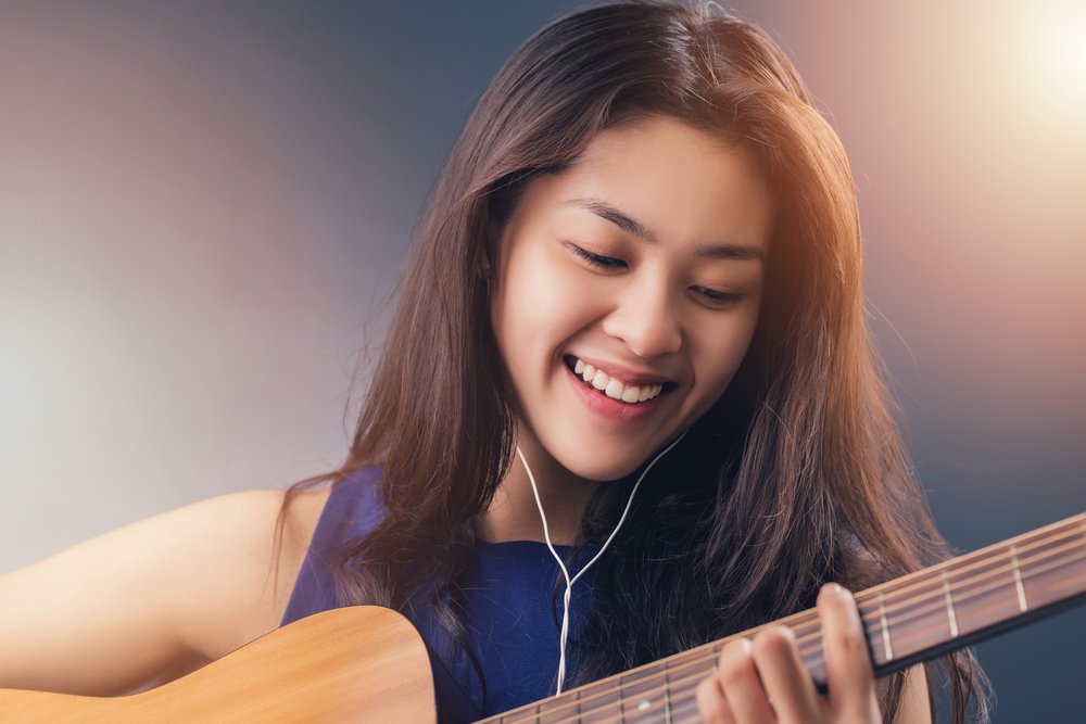 smiling young woman listening to music via ear buds while playing guitar